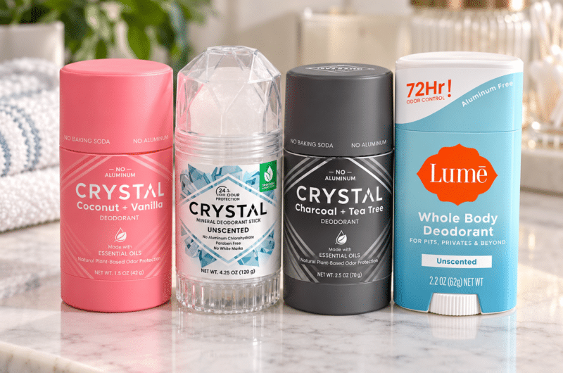 Four natural deodorants from Crystal and Lume displayed on a marble bathroom vanity with greenery, coconut, and vanilla accents.